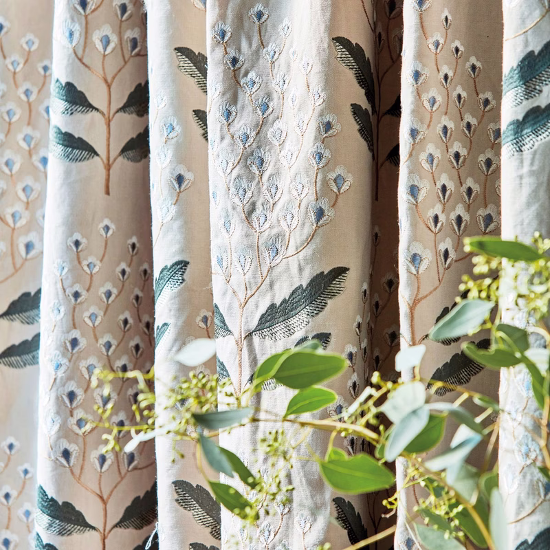Patterned fabric shown as full-length curtains in a room, with a botanical embroidery design and greenery in the foreground.