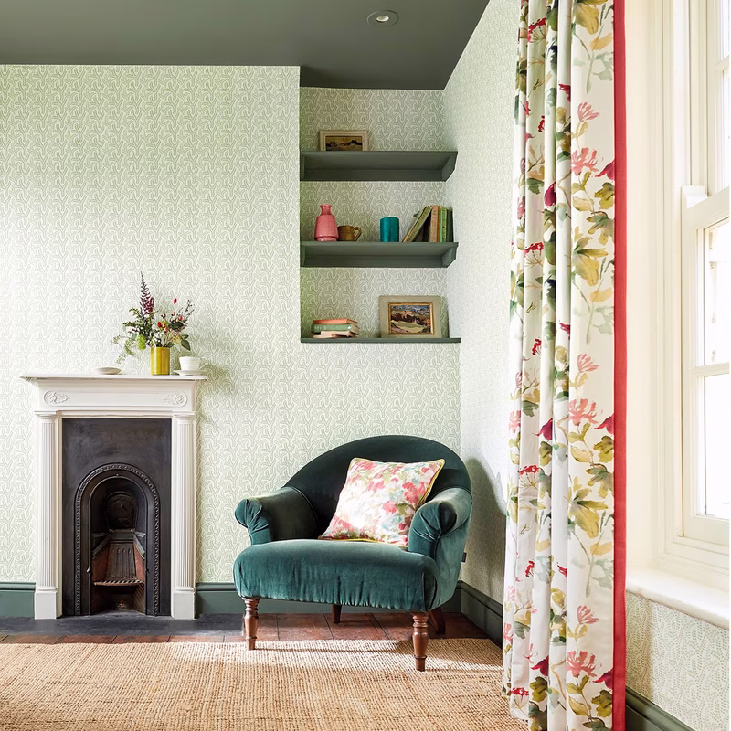A sitting room with patterned wallpaper and a green velvet armchair by a fireplace, with Honey Flowers made up as curtains and a matching cushion.