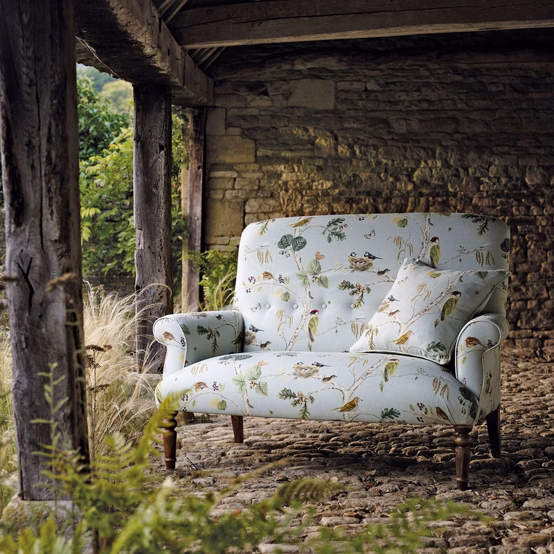Stone barn interior with a floral bird-print armchair upholstered in Woodland Chorus, with a matching cushion.