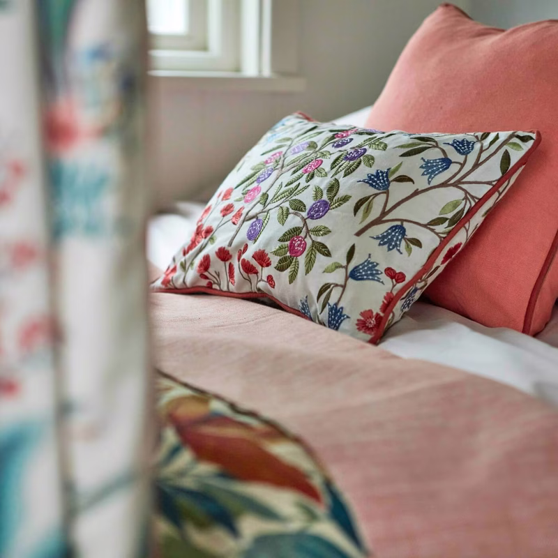 In a bedroom, the fabric is shown as a floral scatter cushion on a bed with a coral cushion behind it.