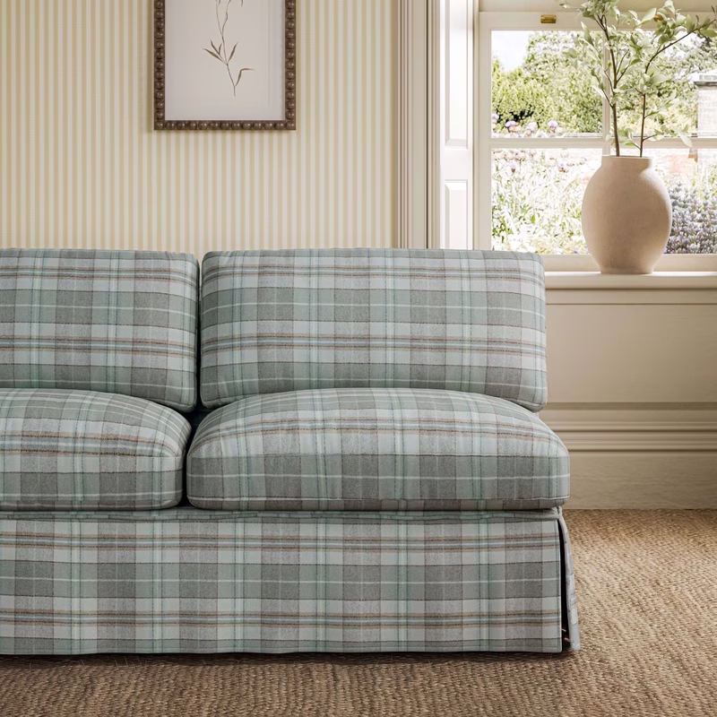 A living room with a sofa upholstered in pale green and grey plaid fabric beside a window and vase of flowers.