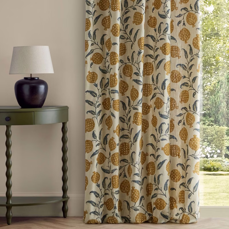 Curtains in a room with a side table and lamp, made up as full-length curtains with a patterned fabric of pomegranates and foliage.