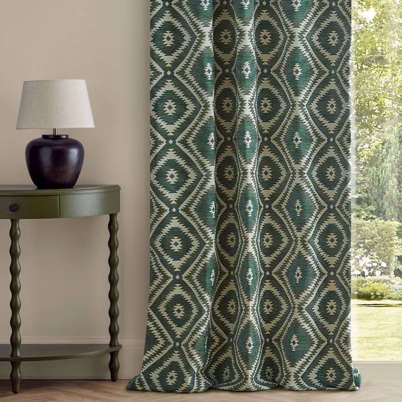 A patterned green and cream curtain hangs by a window beside a table with a lamp in a room.