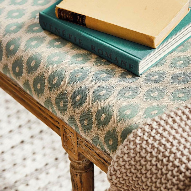Patterned fabric upholstered on a table bench beside books and a knitted cushion.