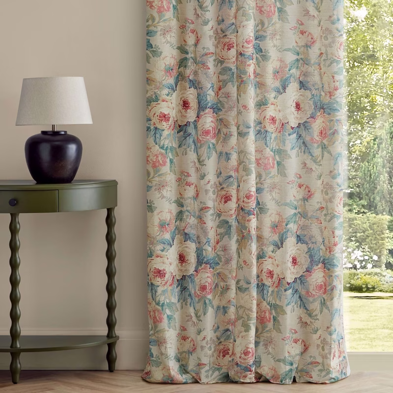 A room with floral patterned curtains hanging beside a window, shown with a table and lamp.