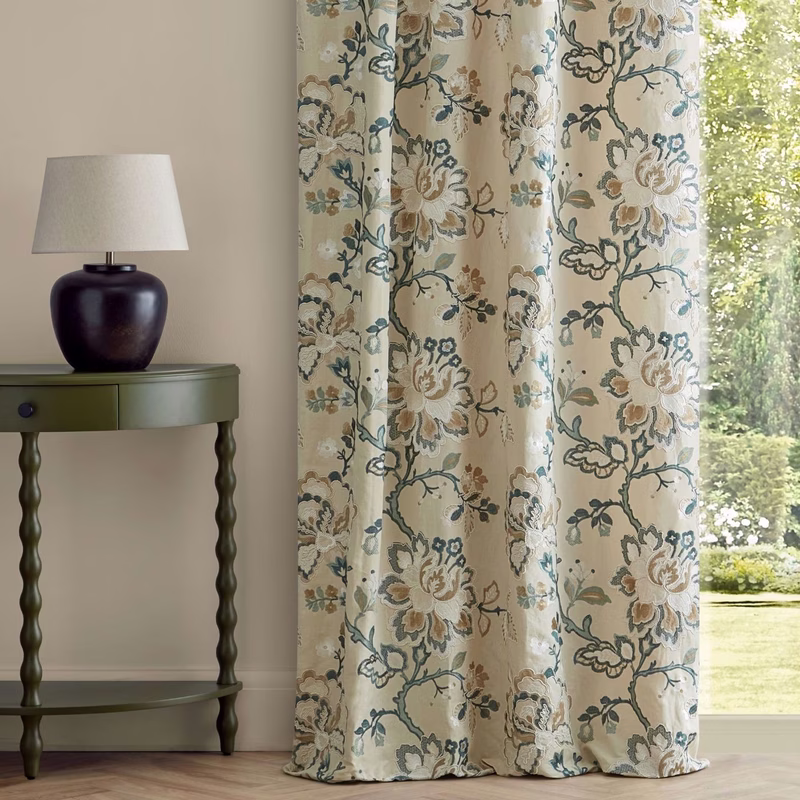 A room with floral patterned curtains hanging at a window beside a lamp on a green table.