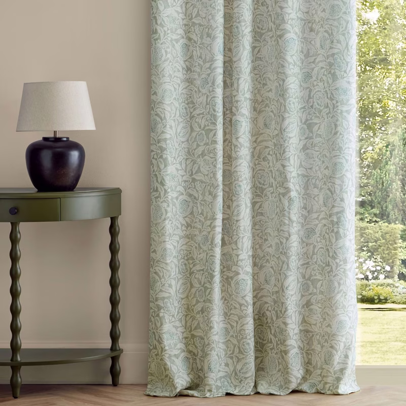 A room with patterned curtains hanging floor to ceiling beside a window and a side table with a lamp.