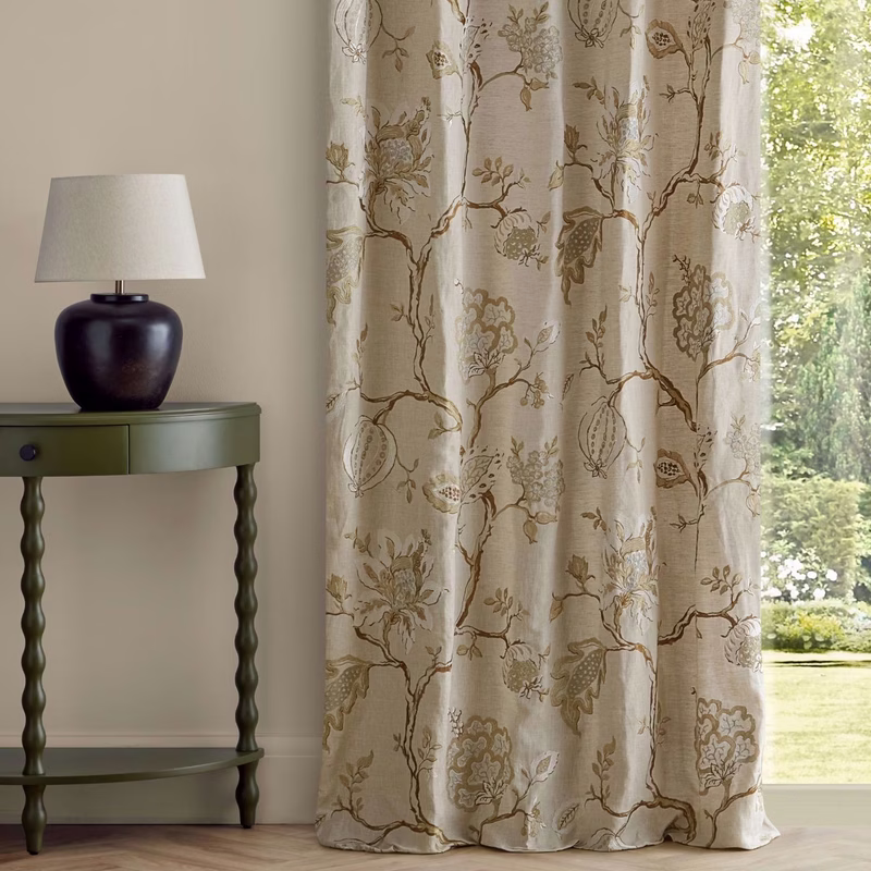 A room with a table lamp beside a window, showing embroidered patterned curtains in a floor-length drape.