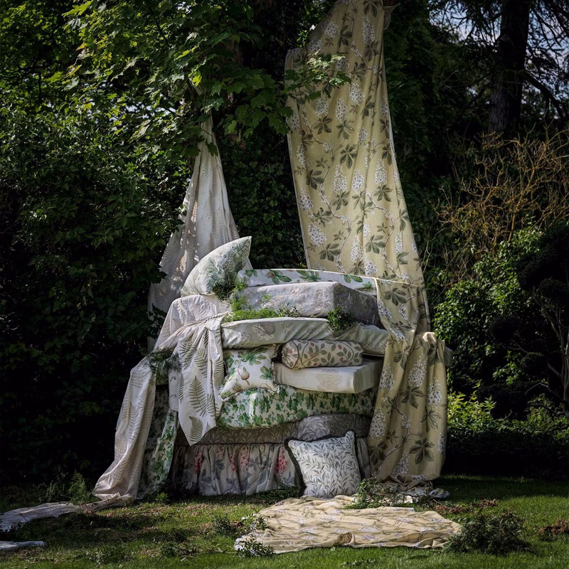 Outdoor scene with draped fabric panels and stacked upholstered cushions showing a fern-print textile, plus a matching loose cloth on the grass.
