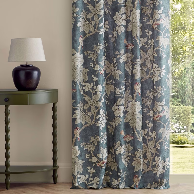 A room with a table lamp and small green side table beside patterned curtains in a botanical print hanging to the floor.