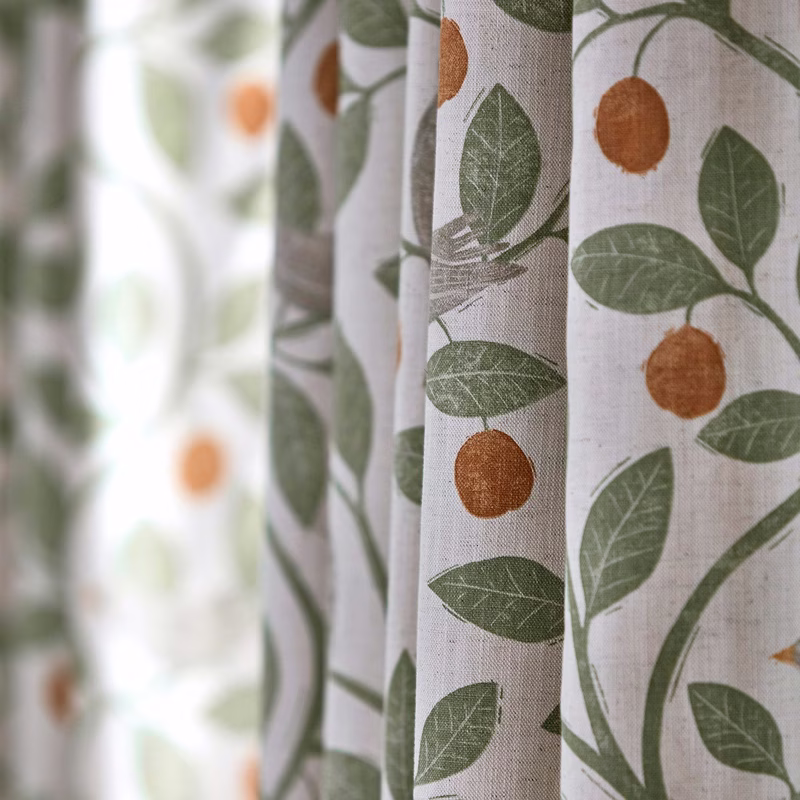 Close-up of patterned fabric made into curtains, with green leaves and orange fruit on a pale background.