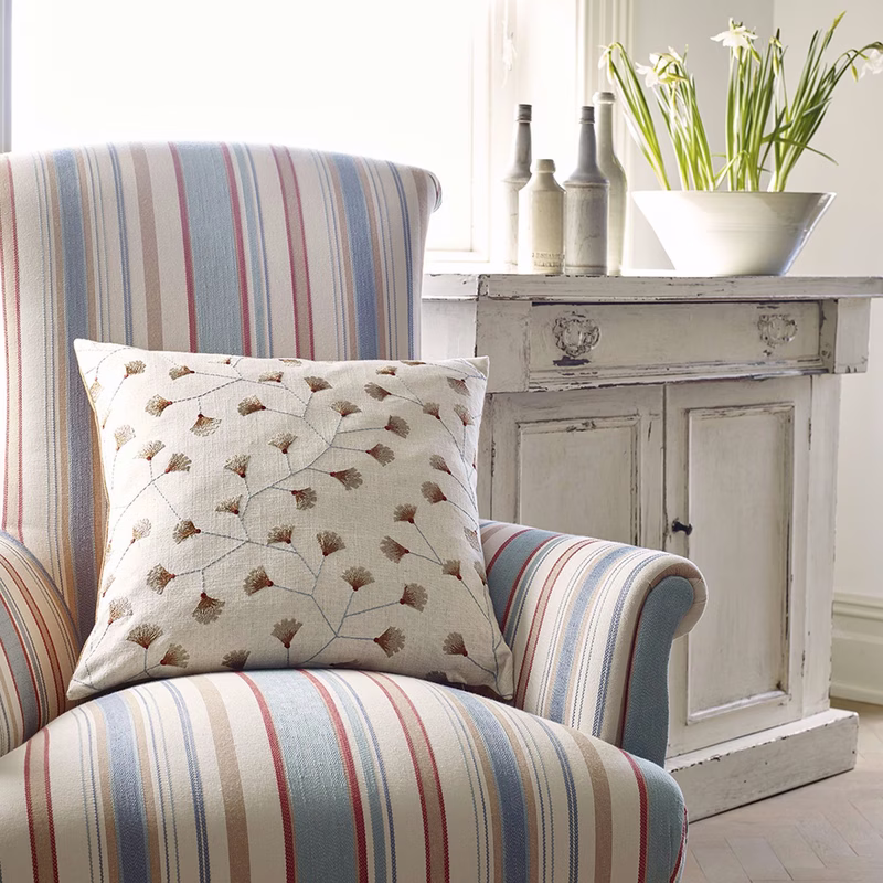 A striped upholstered armchair with a patterned cushion sits beside a painted cabinet topped with vases and flowers.