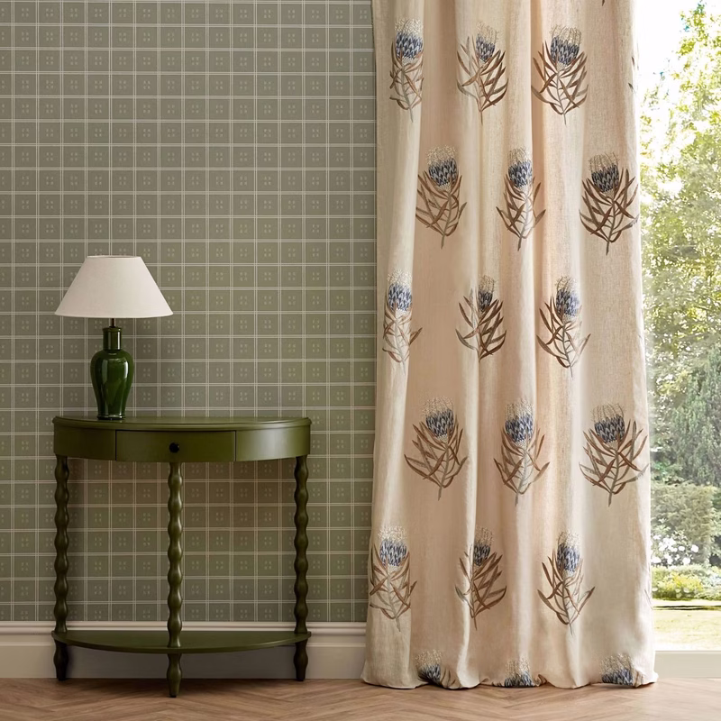 Room with green patterned wallpaper, a lamp and side table, and a cream fabric curtain printed with protea flowers by a window.