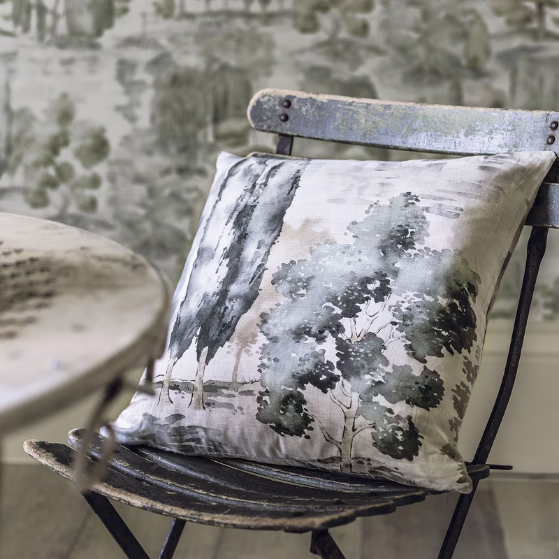 A cushion made from Waterperry fabric on a chair, with a blurred fabric-covered table and wallpapered wall behind.