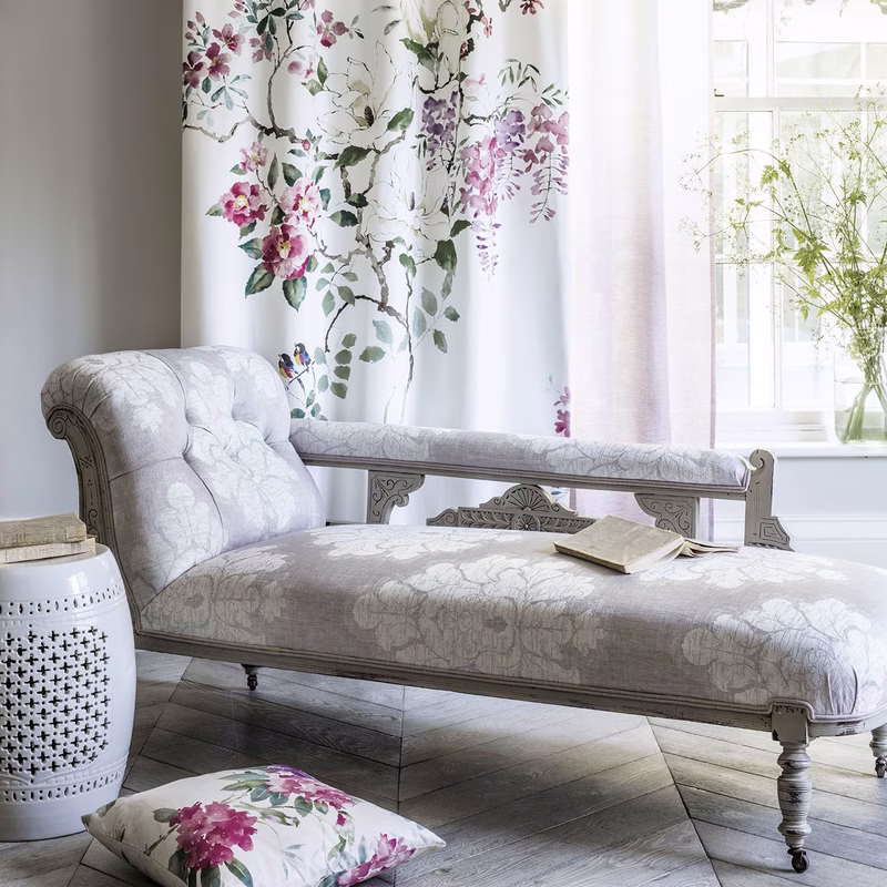 A light room with a chaise longue upholstered in pale floral fabric, with matching curtains and a cushion using the same Magnolia & Blossom design.