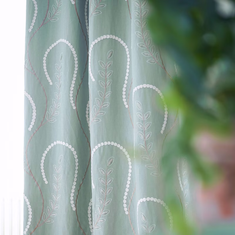 A close-up of patterned green fabric hanging as curtains, with a sheer panel in front and blurred foliage at the right edge.
