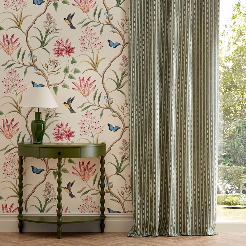 A room with floral bird wallpaper, a green side table and lamp, and floor-length patterned curtains beside a window.
