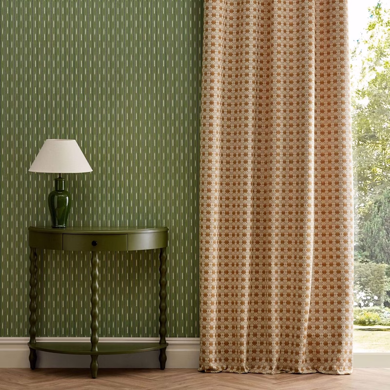 A room with green patterned wall covering, a small table and lamp, and patterned curtains hung beside a window.