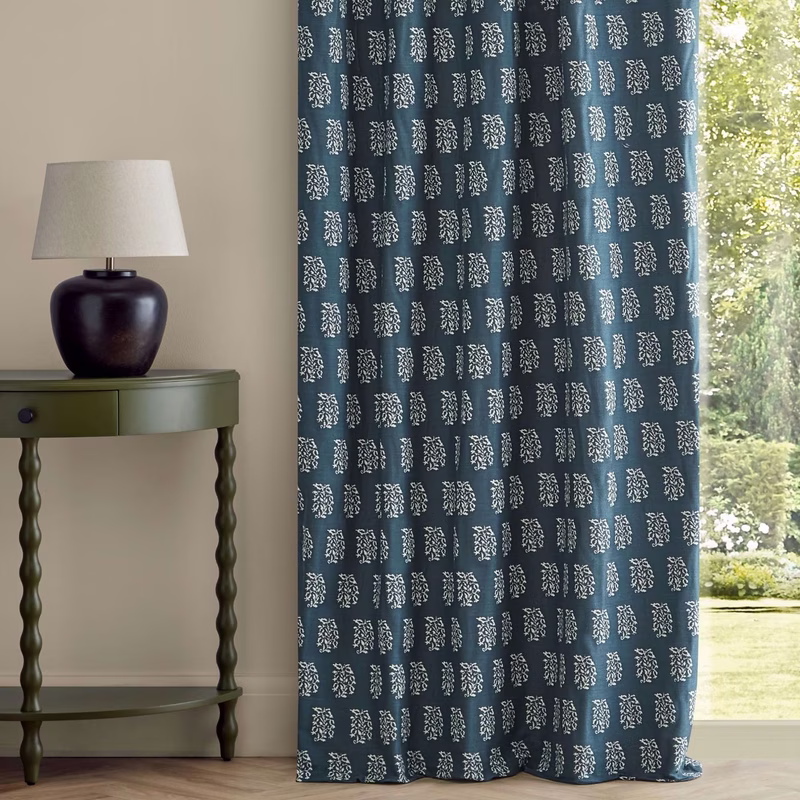 A room with a green side table and lamp beside blue patterned curtains hanging floor length by a window.