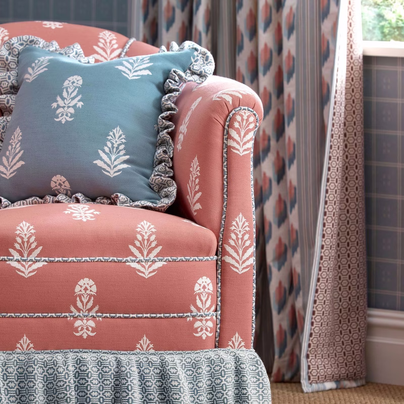 A patterned armchair upholstered in pink floral fabric with a blue ruffled cushion, shown beside patterned curtains and a window.