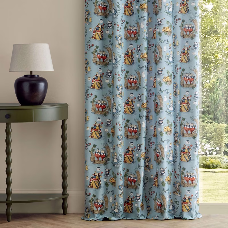 A room with a side table and lamp beside Alice in Wonderland patterned curtains hung with soft folds at a window.