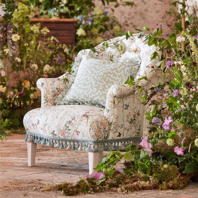 Armchair upholstered in floral Sanderson Bambi fabric with a pale patterned cushion, set among flowers and foliage.