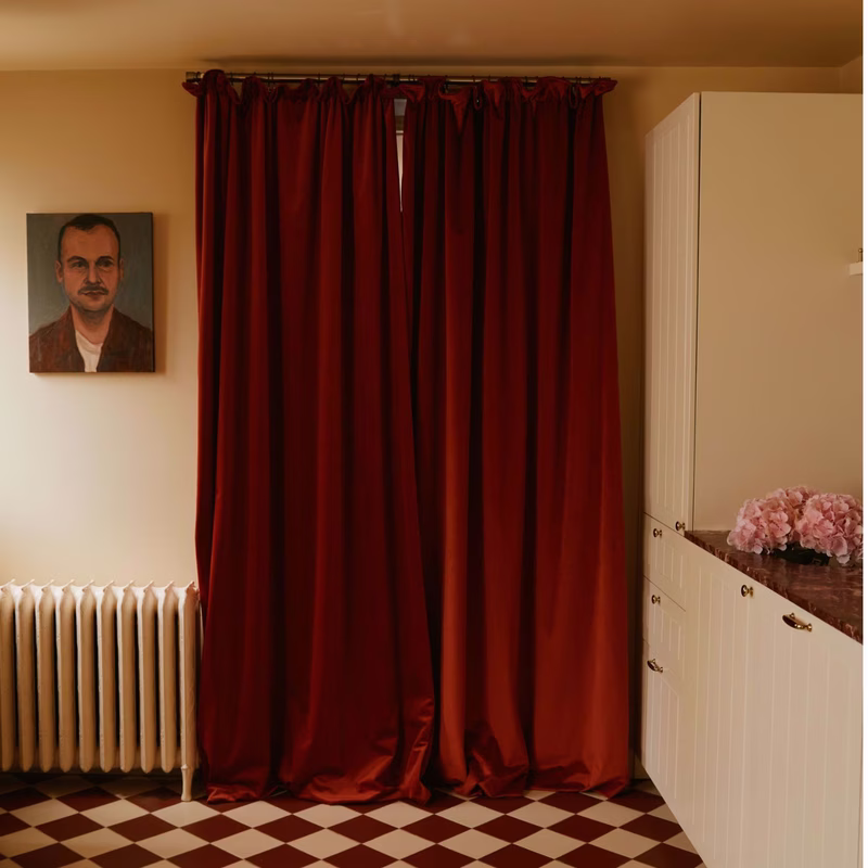 A room with deep red velvet curtains hanging on a track over a doorway or window, with a radiator, portrait, cabinet and tiled floor visible.