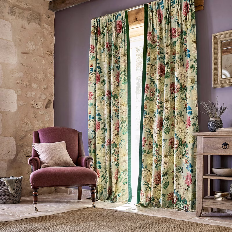 A rustic sitting room with floral patterned curtains hanging at a window, made with a central opening and green leading edges.