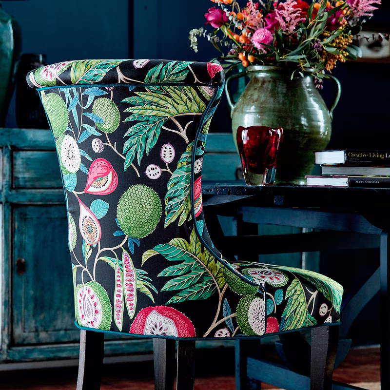 A close-up of a single upholstered dining chair in a room, covered in a dark botanical fruit-print fabric.