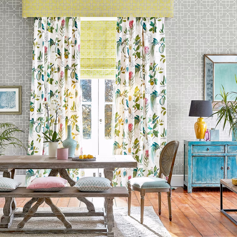 Bright dining room with floral curtains framing a window and a patterned Roman blind above, with matching wall covering and cushions visible.