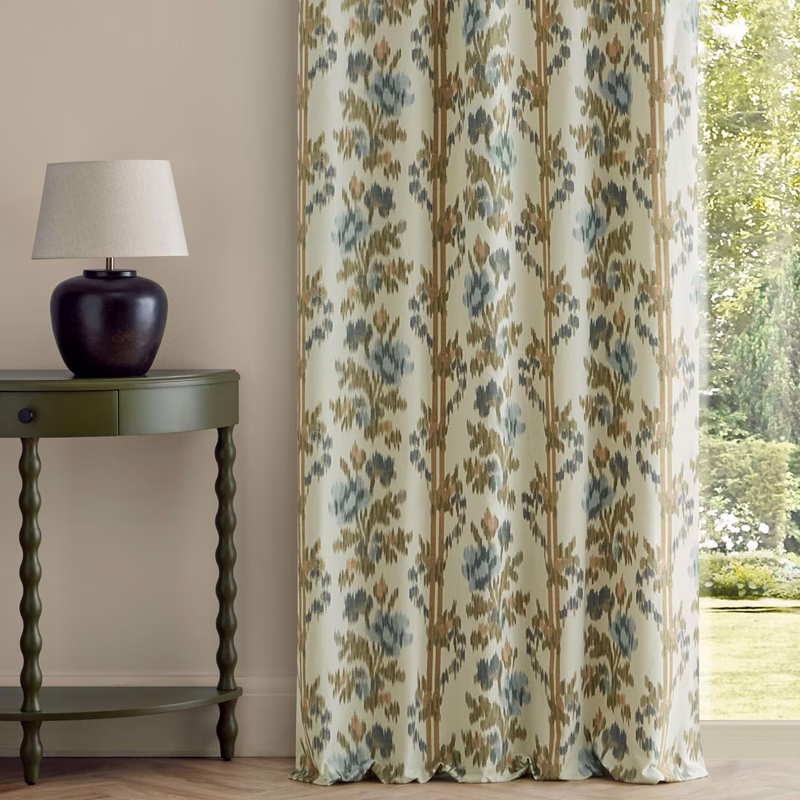 A room with a side table and lamp beside floral patterned curtains hanging floor to ceiling by a window.