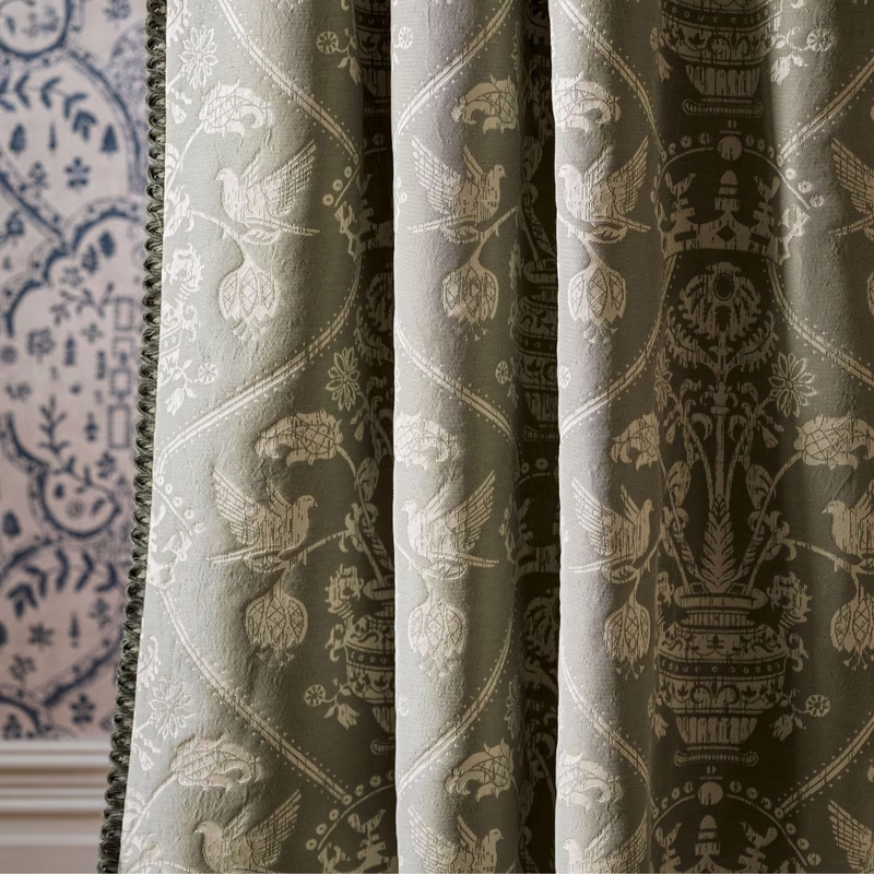 Curtains in a room, showing olive-green patterned fabric with a damask-style bird and urn design hanging in soft folds.
