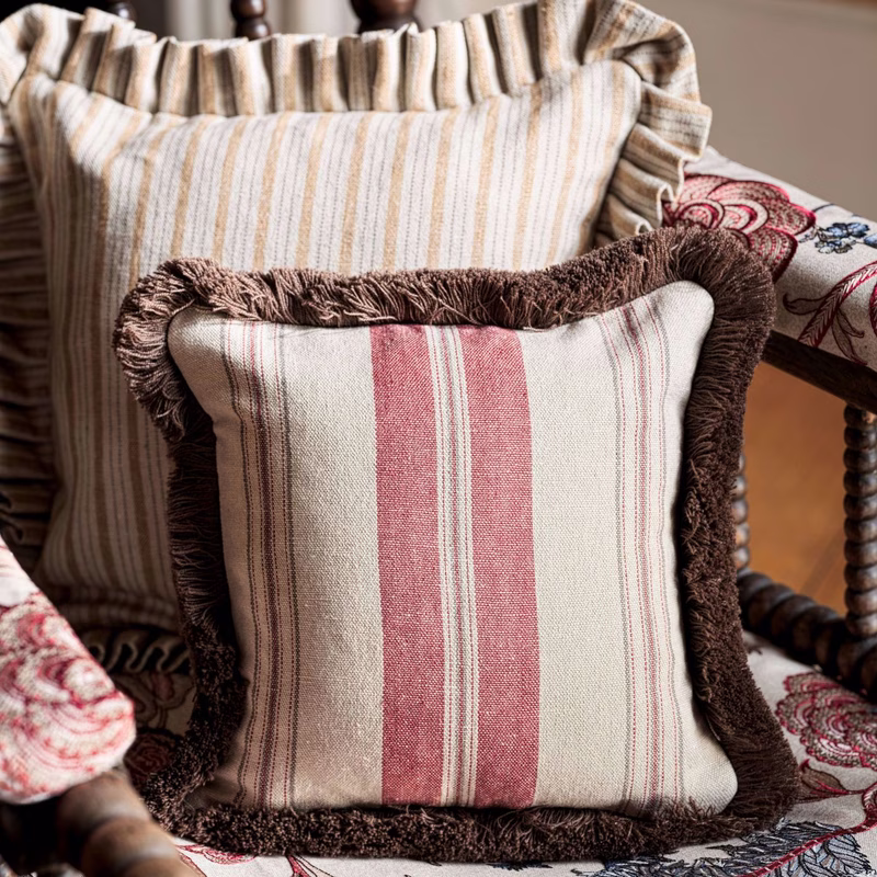 A pair of striped and patterned cushions on a wooden chair, with the Tetbury Stripe fabric made up as a cushion cover with fringe trim.