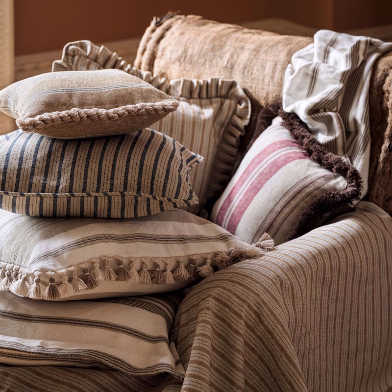 A sofa piled with striped fabric cushions and throws in a warm room, showing the cloth made up as cushions and a draped throw.