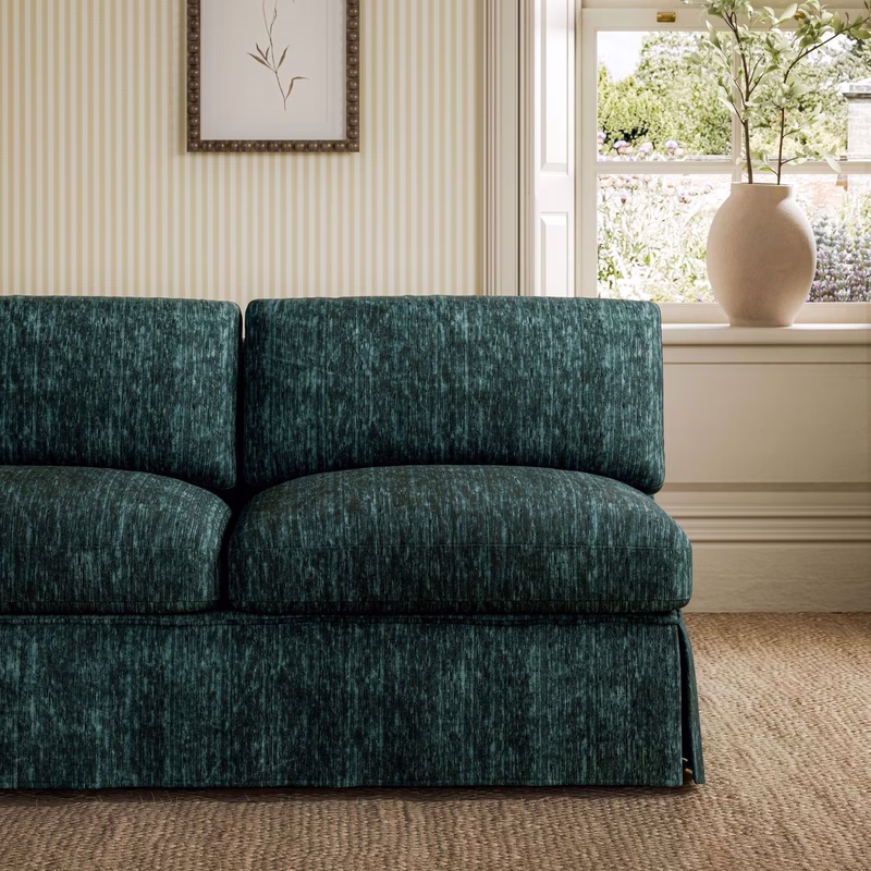 Sofa in a striped cream room upholstered in dark teal velvet with a large window and vase of branches behind it.