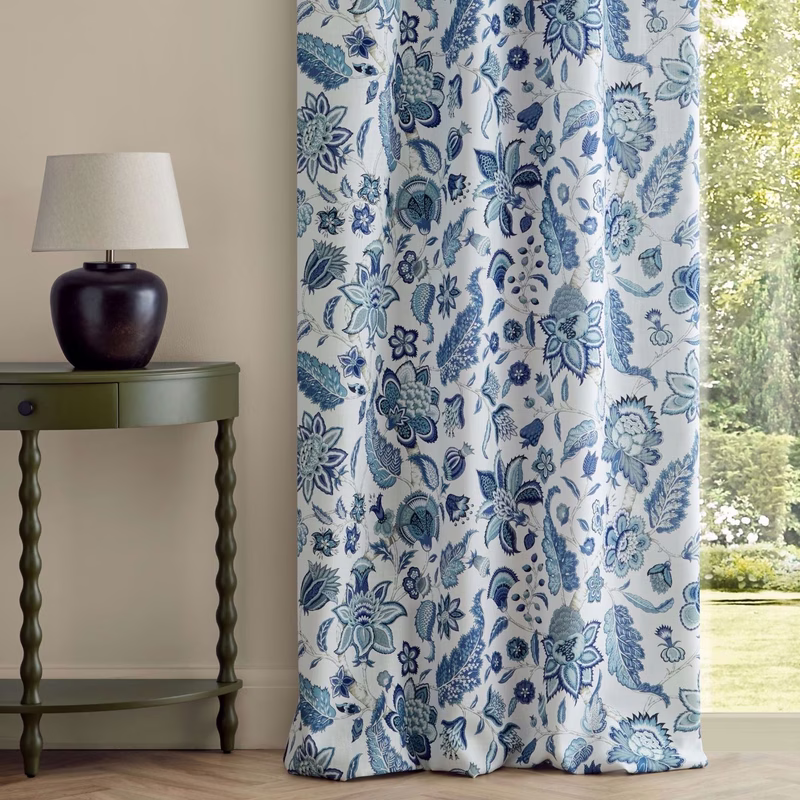A room with a lamp on a small table beside patterned curtain panels in blue and white at a window.