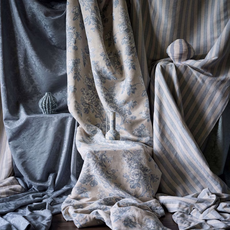 Three draped lengths of fabric in a room setting: a damask, a floral print and a striped cloth, with matching cushions and a small vase on the floor.