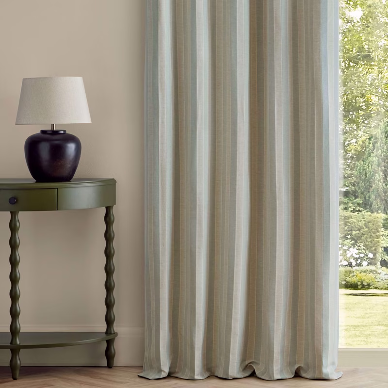 Striped curtains in a room with a green side table and lamp beside a window showing a garden.