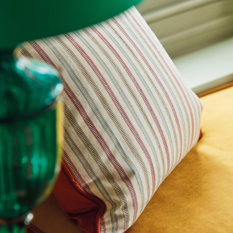 A striped fabric cushion with contrast piping sits on a yellow upholstered seat beside a green glass object.