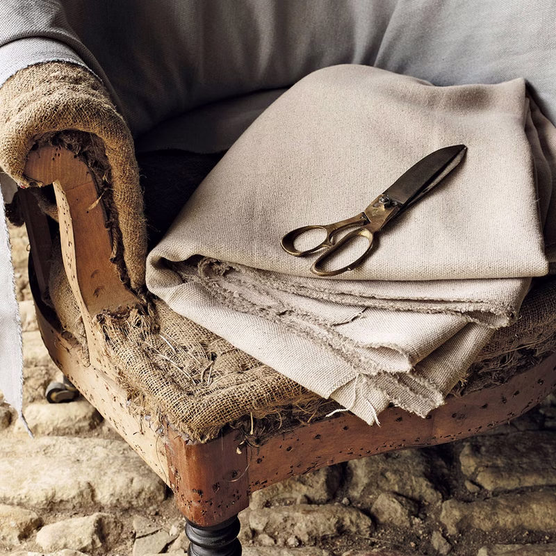 Upholstery workshop scene with folded fabric and scissors resting on an upholstered chair frame with exposed burlap and wood.