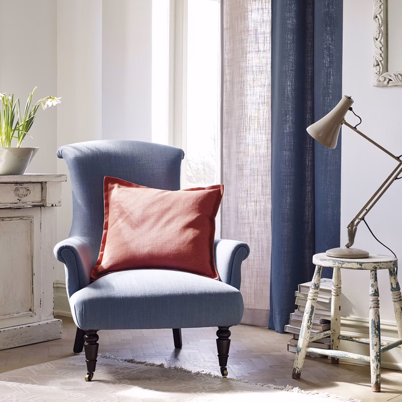 A light room with a blue armchair, cushions and floor-length curtains made from the fabric, plus a small stool and lamp.