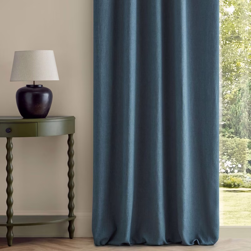 A room with a teal-blue curtain hanging in front of a window beside a green side table and lamp.