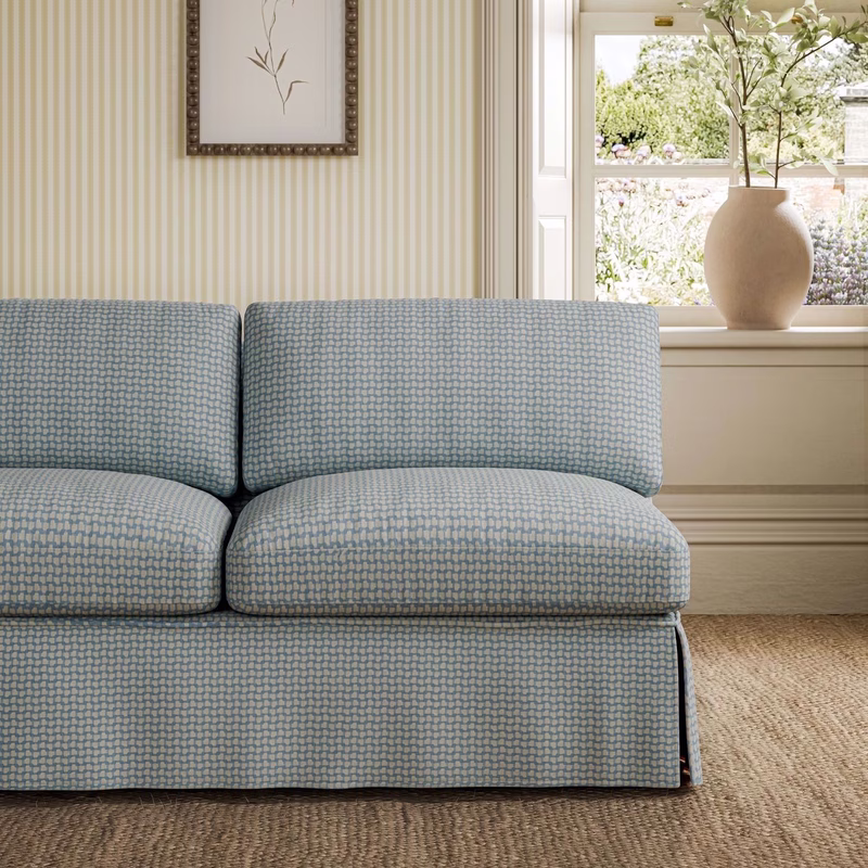 A light room with striped wallpaper, a window and vase, showing a sofa upholstered in blue patterned fabric.