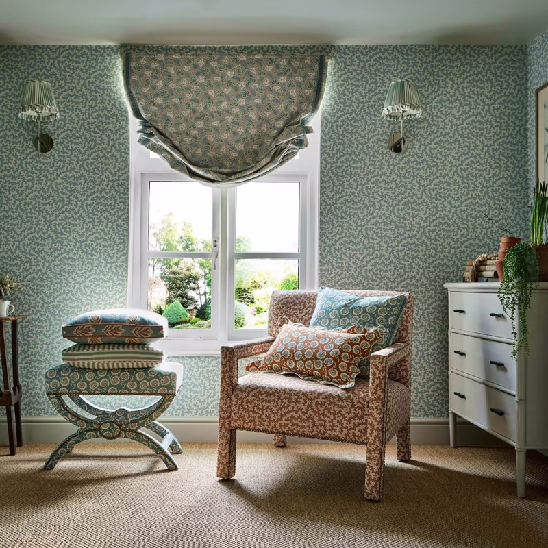 A small room with matching patterned wallpaper, a gathered window blind, an upholstered armchair, and patterned cushions.