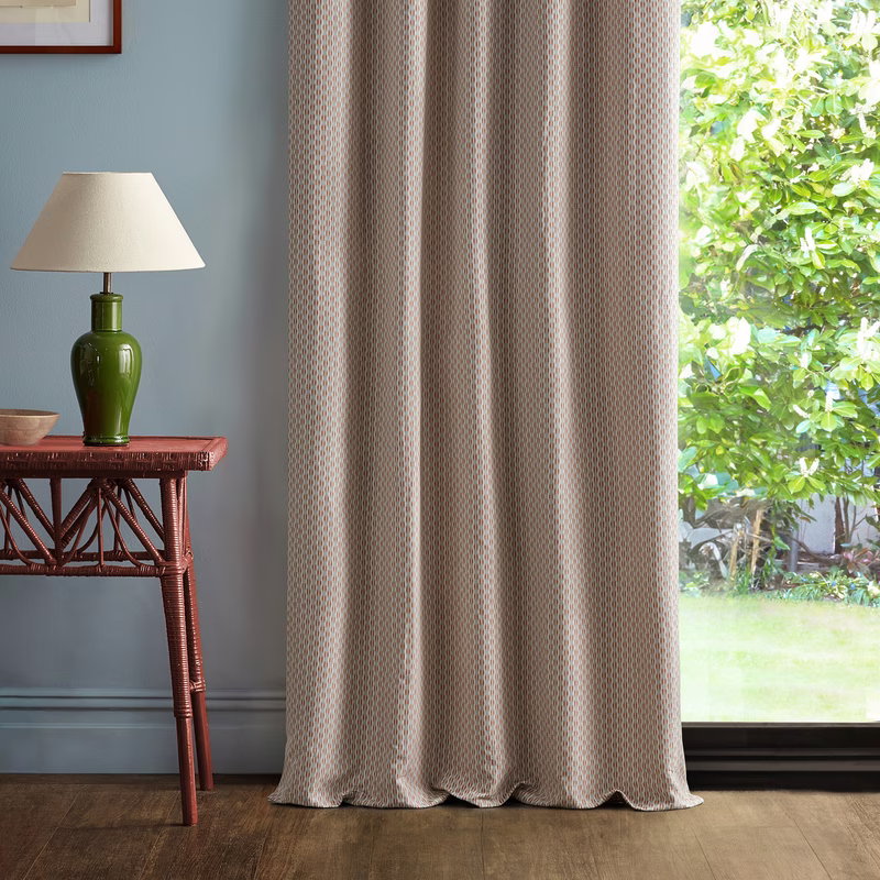 A room with a patterned curtain hanging full length beside a window, with a table and lamp to the left.
