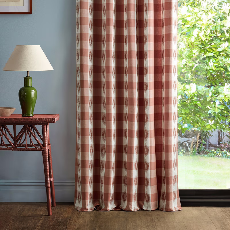 A living room corner with this checked fabric made up as full-length curtains beside a window, with a table lamp and side table at left.