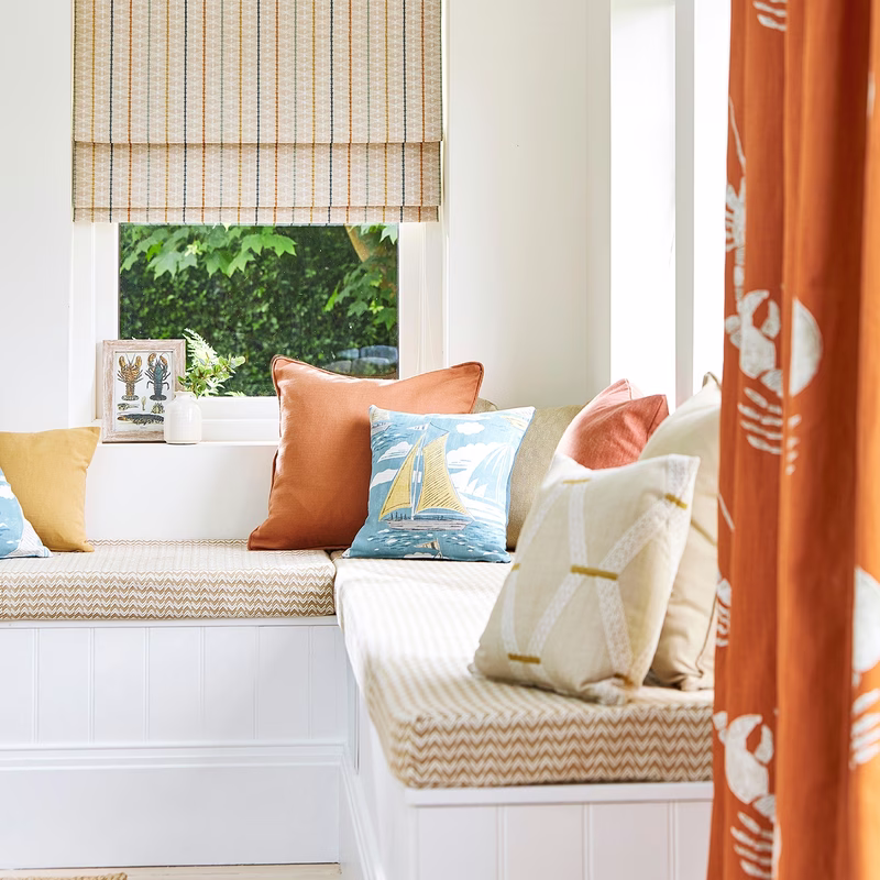 Window seat with patterned cushions and a tan check Roman blind, plus orange curtains at the side.
