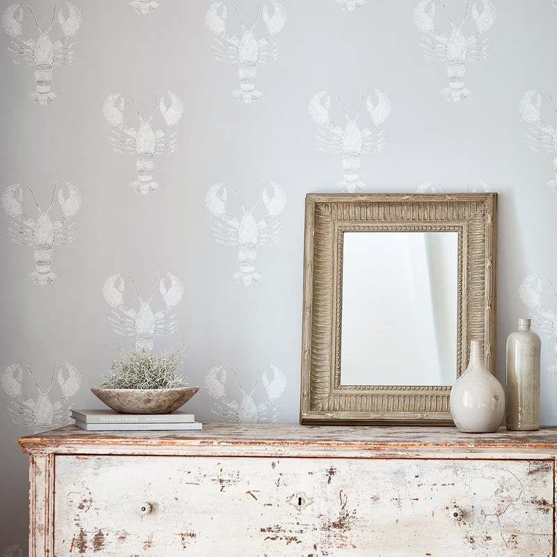 A room with pale lobster-pattern wallpaper behind a distressed sideboard, mirror and ceramics.