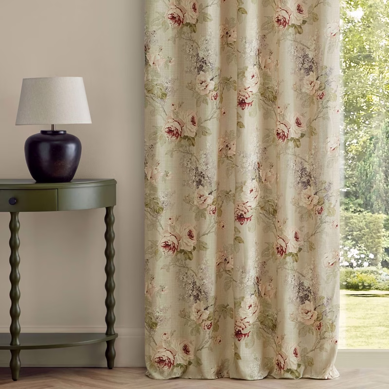 A room with a floral patterned curtain panel hanging at a window beside a small table and lamp.