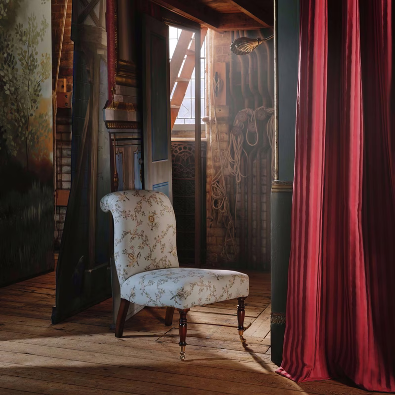An upholstered chair in a rustic room, covered in a floral fabric and shown beside hanging striped curtains.